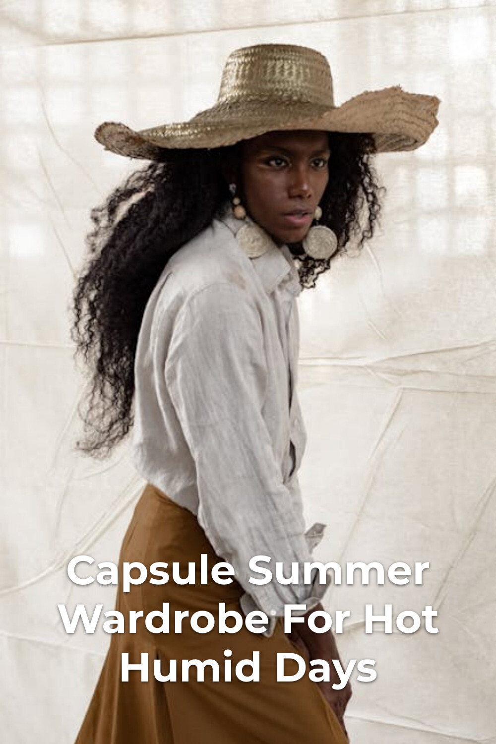 Capsule summer wardrobe look with straw hat, light shirt and brown skirt against a soft fabric backdrop
