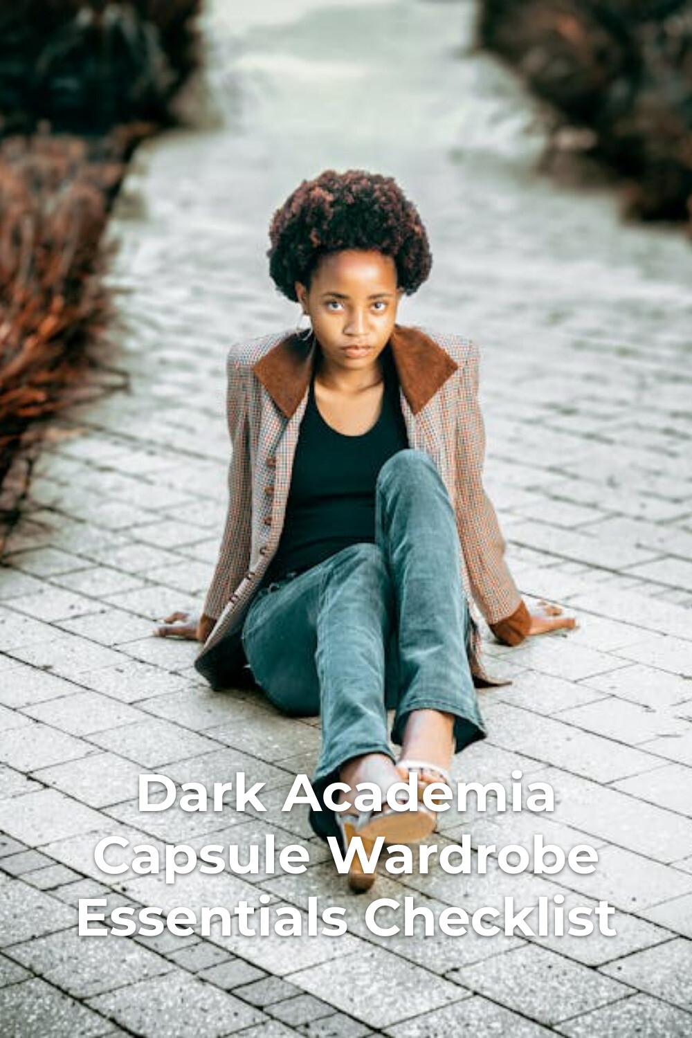 Dark academia capsule wardrobe look with jacket, black top, jeans and sandals, seated on a paved path by bushes