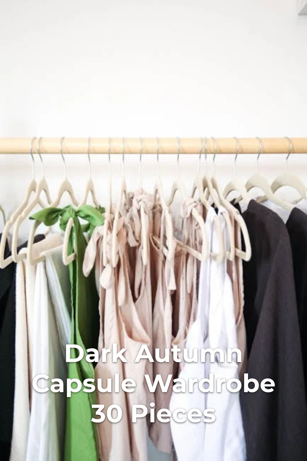 Dark autumn capsule wardrobe with light-toned clothes and a dark jacket on wooden hangers against a white wall
